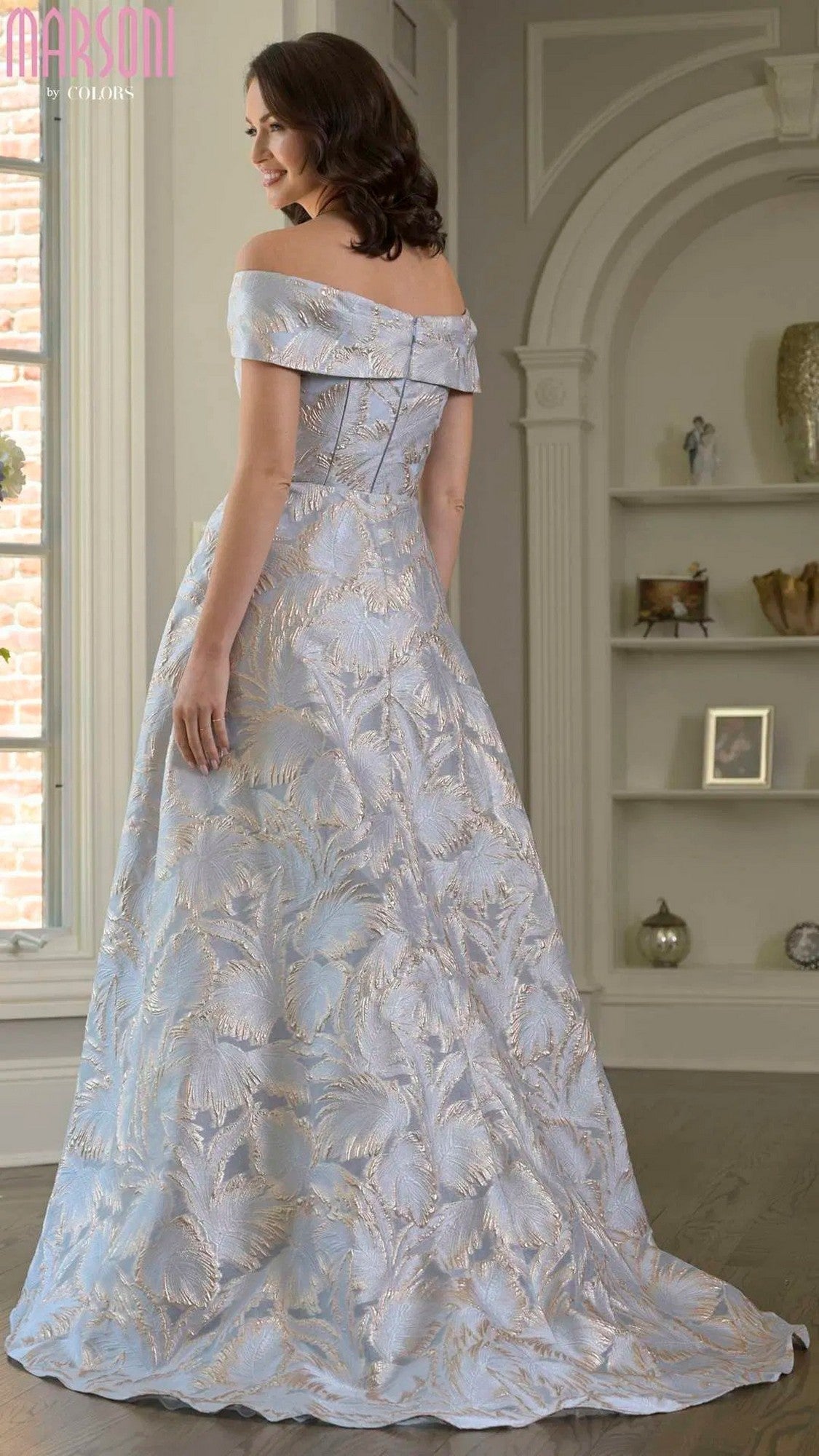 A woman wearing the Marsoni MV1382 long formal dress in light blue with a floral pattern stands in a well-lit, elegant room, facing away from the camera.